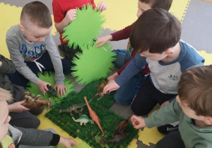 Widok na grupę chłopców, którzy układają prehistoryczny las wykorzystując plastikowe dinozaury, zielone serwetki, trawki.